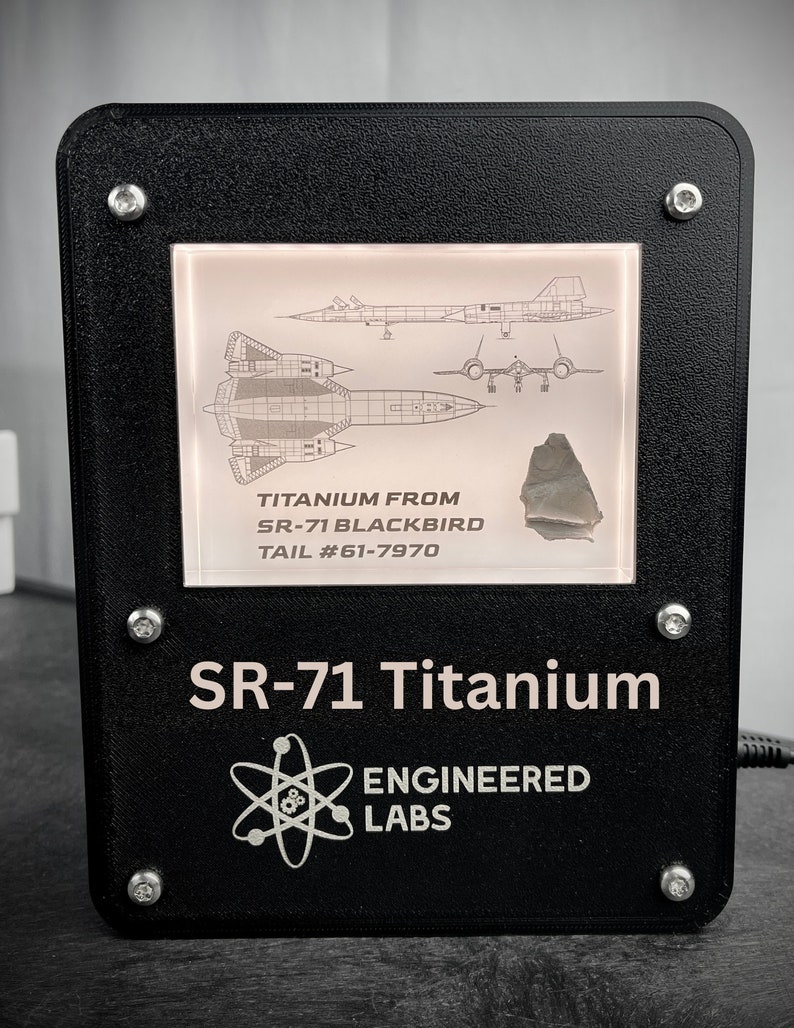 Puede incluir: Una placa de exhibici&oacute;n negra con un marco plateado que muestra un trozo de titanio de un avi&oacute;n SR-71 Blackbird. La placa presenta un diagrama del SR-71 y el texto "TITANIUM FROM SR-71 BLACKBIRD TAIL #61-7970". Debajo del marco, la placa dice "SR-71 Titanium" y "ENGINEERED LABS" con un s&iacute;mbolo de &aacute;tomo.