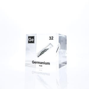 May include: A transparent, rectangular block displaying the element Germanium. The label shows the symbol 'Ge', atomic number 32, and an illustration of Germanium in a test tube. The block is set against a white backdrop.