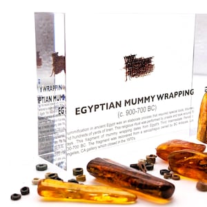 May include: A close-up of a small piece of brown linen fabric in a clear acrylic display case. The case has a label that reads "Egyptian Mummy Wrapping (c. 900-700 BC)". The label explains that mummification in ancient Egypt was an elaborate process that required special tools, hundreds of yards of linen, and took around 70 days. This fragment of mummy wrapping dates from Egypt's Third Intermediate Period (c. 1070-700 BC). The fragment was recovered from a sarcophagus owned by BC Antiques, Los Angeles, CA gallery which closed in the 1970's.