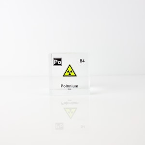 May include: A clear, square block with the element symbol "Po" and the number "84" printed in black. The block features a yellow radiation hazard symbol and the word "Polonium" in black text. The block is reflected on a white surface.