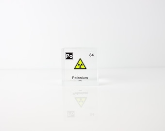 May include: A clear, square block with the element symbol "Po" and the number "84" printed in black. The block features a yellow radiation hazard symbol and the word "Polonium" in black text. The block is reflected on a white surface.