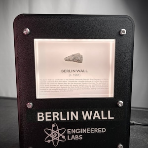 Puede incluir: Una vitrina negra con un peque&ntilde;o trozo del Muro de Berl&iacute;n en su interior. La vitrina tiene un fondo blanco con el texto "BERLIN WALL (c. 1961)" y una descripci&oacute;n de la historia del muro. La parte inferior de la vitrina tiene el texto "BERLIN WALL ENGINEERED LABS" en blanco.