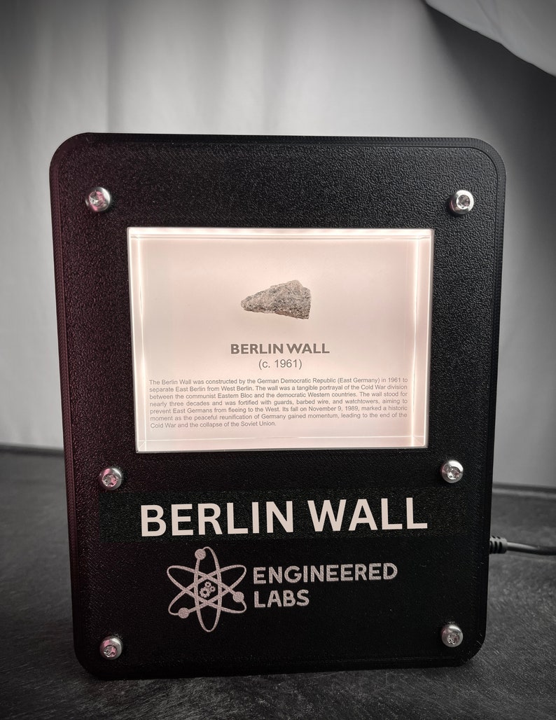 Puede incluir: Una vitrina negra con un peque&ntilde;o trozo del Muro de Berl&iacute;n en su interior. La vitrina tiene un fondo blanco con el texto "BERLIN WALL (c. 1961)" y una descripci&oacute;n de la historia del muro. La parte inferior de la vitrina tiene el texto "BERLIN WALL ENGINEERED LABS" en blanco.