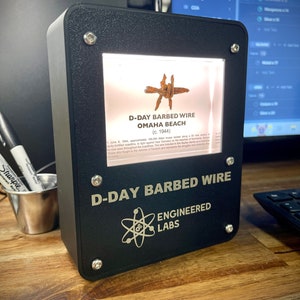 以下が含まれることがあります： オマハビーチ、ノルマンディー、フランスの有刺鉄線の一部が、照明付きの窓から見える黒いディスプレイケース。ケースには、「D-Day Barbed Wire Engineered Labs」という金色の文字が刻まれています。