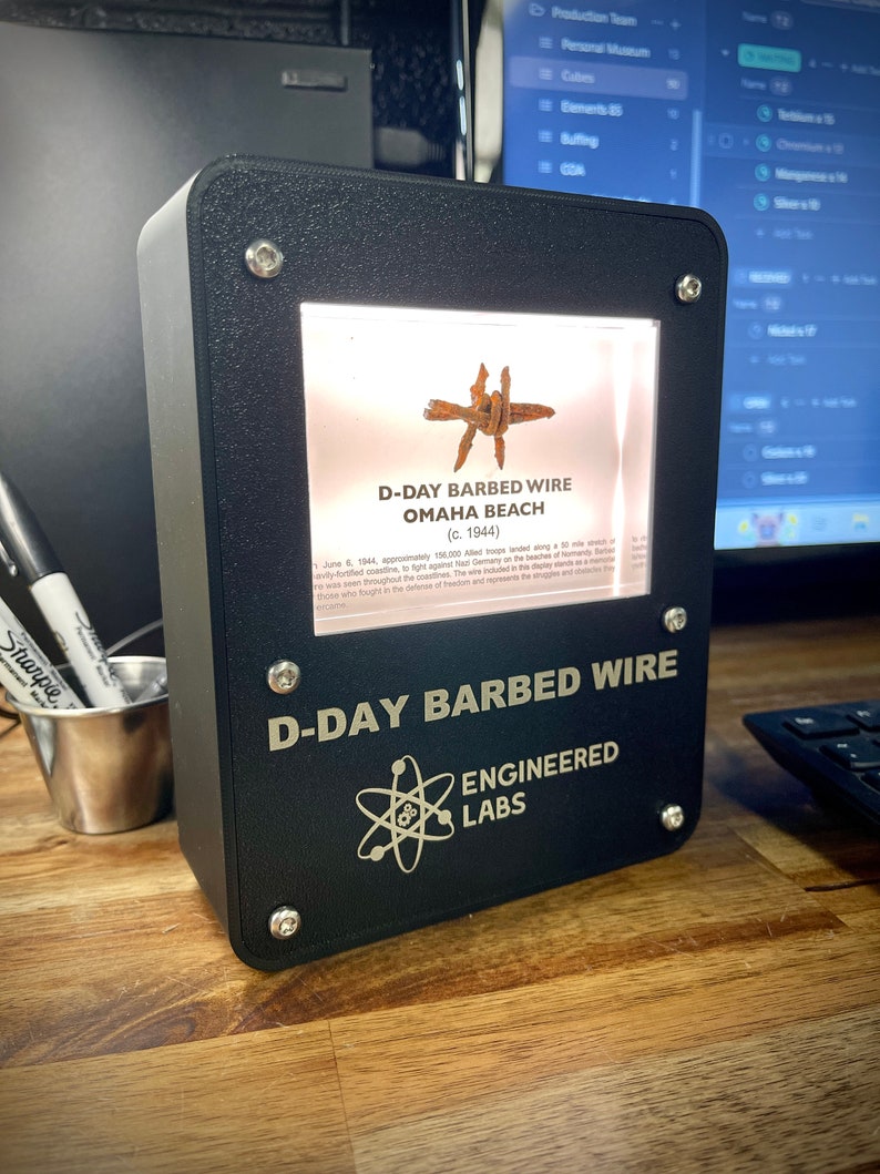 Puede incluir: Una vitrina negra con una ventana iluminada que muestra un trozo de alambre de p&uacute;as de la playa de Omaha, Normand&iacute;a, Francia. La vitrina tiene una inscripci&oacute;n dorada que dice "D-Day Barbed Wire Engineered Labs".