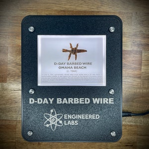 Puede incluir: Un expositor enmarcado en negro con fondo blanco que muestra un trozo de alambre de p&uacute;as de Omaha Beach durante el D&iacute;a D. El texto dice "D-Day Barbed Wire Omaha Beach (c. 1944)". Debajo del expositor est&aacute; el texto "D-Day Barbed Wire Engineered Labs".