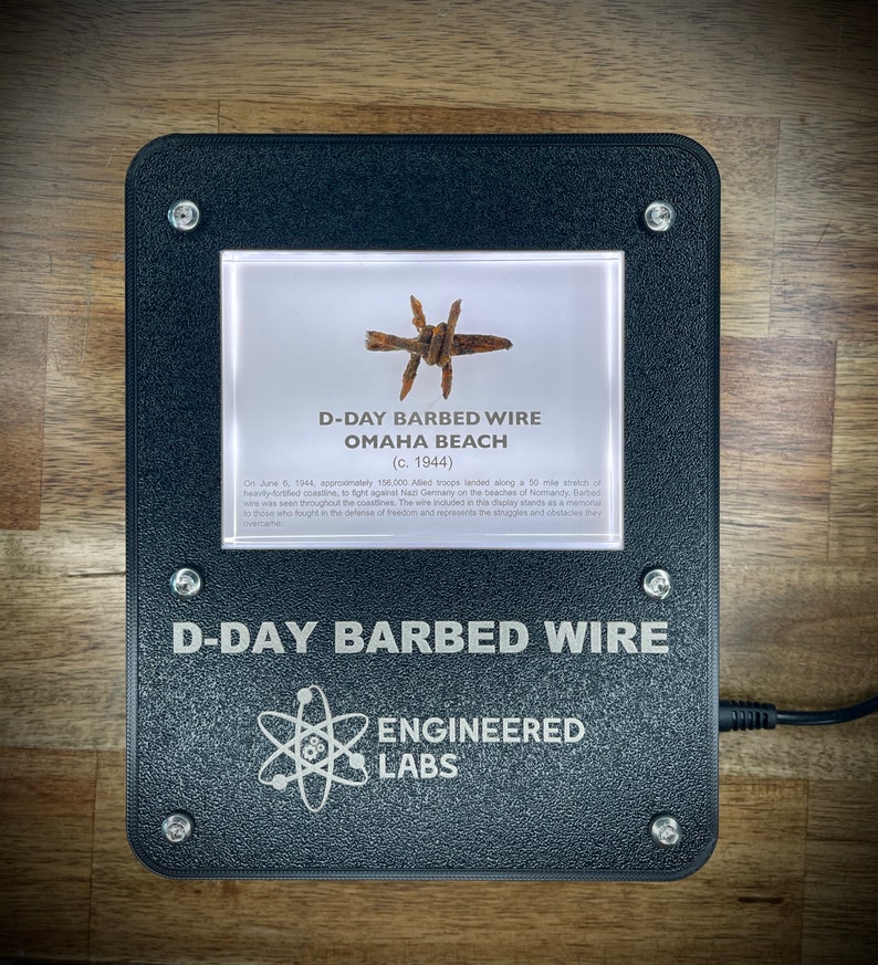 Puede incluir: Un expositor enmarcado en negro con fondo blanco que muestra un trozo de alambre de p&uacute;as de Omaha Beach durante el D&iacute;a D. El texto dice "D-Day Barbed Wire Omaha Beach (c. 1944)". Debajo del expositor est&aacute; el texto "D-Day Barbed Wire Engineered Labs".