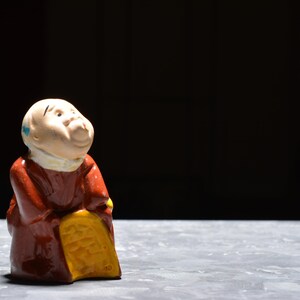 Puede incluir: Figura de cerámica de un anciano sonriente con una túnica marrón, sosteniendo un saco amarillo. La figura tiene la cabeza calva con una mancha azul y una bufanda blanca. El fondo es negro.