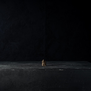 May include: A small, textured, brown object stands on a dark gray, rectangular surface. The background is a solid black, creating a high-contrast, minimalist composition. The object appears to be the focal point.