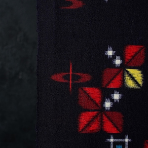 May include: A dark navy blue fabric featuring a repeating pattern of geometric shapes. The design includes red, yellow, and green squares with white cross-stitch accents, alongside red oval shapes with a thin vertical line through the center.