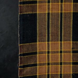 Könnte beinhalten: Ein Stück Stoff mit einem Karomuster aus sich kreuzenden Linien in den Farben Schwarz, Dunkelbraun und Senfgelb. Das Textil weist eine gewebte Struktur mit einer sichtbaren, unversäuberten Kante auf der linken Seite des Karomusters auf.