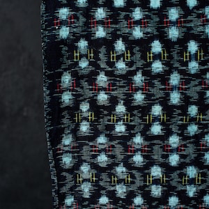 Puede incluir: Una tela oscura con un patrón geométrico repetitivo en azul claro, con pequeños acentos verticales en rojo y amarillo. El tejido muestra una textura tejida con una disposición en cuadrícula consistente de formas en toda la superficie.