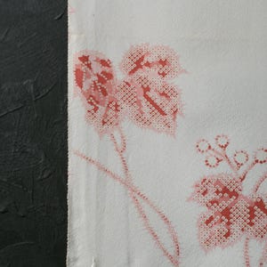 Puede incluir: Tela blanca con un patrón repetido de hojas y vides rojas. Las hojas tienen un diseño detallado y punteado. La tela está sobre un fondo oscuro y texturizado, creando un contraste.