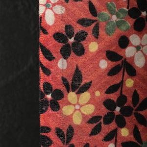 Puede incluir: Primer plano de una tela floral con fondo rojo. La tela presenta flores negras, blancas y amarillas, junto con hojas verdes y lunares blancos. Una tela negra está en el lado izquierdo.