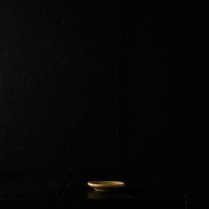 May include: A small, round, ceramic dish with a slightly raised edge. The dish is a muted gold color and sits on a dark, textured surface. The background is a solid black, creating a dramatic contrast and highlighting the dish.