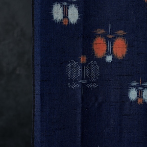May include: A deep navy blue fabric featuring a repeating ikat-style pattern of stylised floral or lantern motifs in shades of bright orange, soft white, and light blue, with a visible slubbed texture throughout the woven material surface.