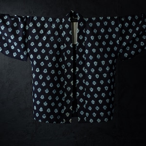 Peut inclure: Une veste haori bleu marine foncé ornée d'un motif répétitif de fleurs bleu clair en forme de losange. Le vêtement présente des manches larges trois-quarts, une ouverture frontale droite et un col structuré, présenté sur un cintre.