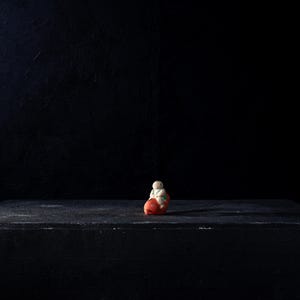 Puede incluir: Pequeña figura decorativa de una figura sentada con cuerpo blanco, base roja y detalles verdes. La figura está colocada sobre una superficie oscura y texturizada sobre un fondo negro.