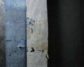 May include: Textile art piece with a distressed aesthetic. The artwork is divided into two vertical panels, one blue and one off-white, both showing signs of wear and tear, including holes and peeling paint. The piece hangs from a metal rod.