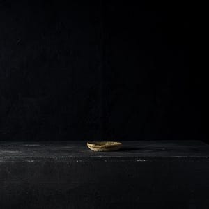 May include: A small, round, textured bowl with a light brown color sits on a dark, rectangular surface. The background is a solid black. The bowl appears to be made of a natural material, possibly clay or stone.