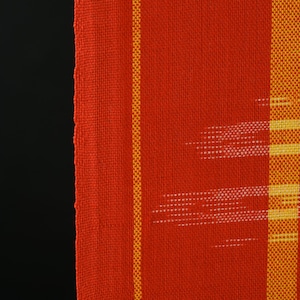 Könnte beinhalten: Nahaufnahme eines gewebten Textils mit leuchtend roter Grundfarbe. Vertikale gelbe Streifen und weiße lineare Muster setzen Kontraste. Der Stoff hat ein strukturiertes Aussehen, was auf eine handgefertigte oder handwerkliche Qualität hindeutet. Der Hintergrund ist tiefschwarz.