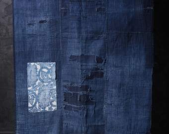 May include: A large, indigo-dyed textile with visible patches and distressed areas. A smaller rectangular patch with a white floral pattern is attached. The fabric has a textured appearance, suggesting a vintage or handcrafted origin.