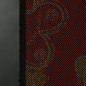 May include: Close-up of a woven textile with a repeating pattern. The fabric features a dark red background with a grid of small squares. White and yellow shapes are woven into the fabric. A black border runs along the left edge.