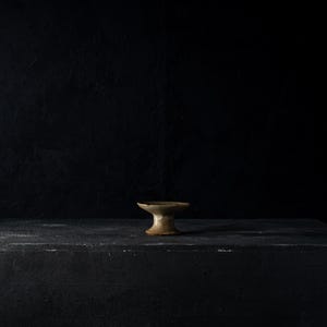 May include: A small, antique-style pedestal bowl with a wide, shallow top and a short, flared base. The bowl is a muted tan color, and it sits on a dark, textured surface against a black backdrop.