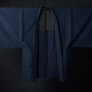May include: A dark blue indigo-dyed haori jacket featuring a subtle, fine grid pattern across the fabric. The garment has wide, straight sleeves, a structured collar, and a contrasting dark grey inner lining visible at the neck and upper back area.