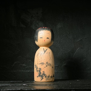 Könnte beinhalten: Eine Kokeshi-Puppe aus Holz mit schwarz lackierter Frisur und geschlossenen Augen. Die Puppe hat einen hellen Holzkörper mit einem bemalten Landschaftsdesign in Schwarz. Die Puppe steht auf einer dunklen Oberfläche vor einem schwarzen Hintergrund.