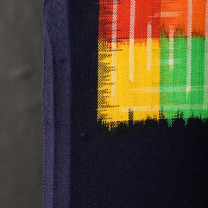May include: Close-up of a section of fabric with a vibrant, abstract design. The design features blocks of red, orange, yellow, and green, with white vertical lines. The fabric has a dark blue border.