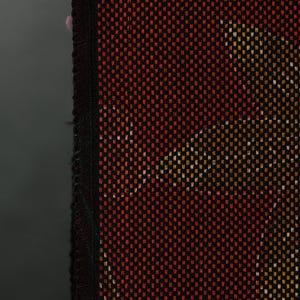 Op de afbeelding: Close-up van een geweven textiel met een herhalend patroon. De stof is voornamelijk zwart met een raster van kleine rode vierkantjes. Accentkleuren van geel en wit creëren een subtiel ontwerp. De rand is afgewerkt met een zwarte rand.