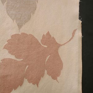 Może przedstawiać: Zbliżenie tkaniny z powtarzającym się wzorem liści. Liście mają stonowany różowy kolor na kremowym tle. Tkanina ma teksturowany wygląd, a krawędź tkaniny jest widoczna.