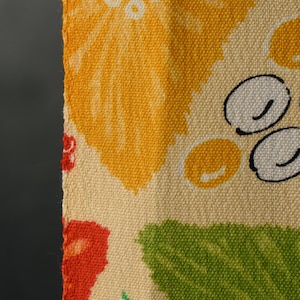 May include: Close-up of a woven fabric with a floral pattern. The design features large yellow and orange flowers, green leaves, and white and black circular shapes on a cream background. The fabric has a textured appearance.
