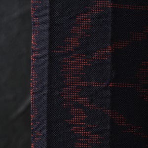 May include: Close-up of a textured fabric with a dark navy blue background and a repeating pattern in a reddish-orange color. The pattern has a zig-zag design. The fabric is likely used for clothing or home decor.