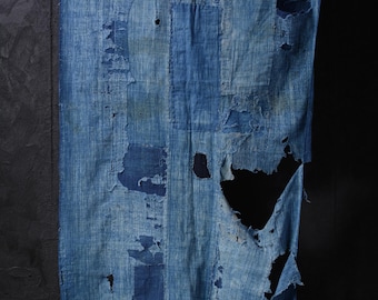 May include: A distressed indigo textile with visible patches and tears. The fabric has a vertical orientation, with varying shades of blue. The design features visible mends and repairs, showcasing a rustic aesthetic. The textile is likely a decorative piece or a repurposed garment.