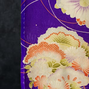 May include: A vibrant purple fabric featuring a detailed floral pattern with large white and pale yellow chrysanthemum-like blossoms, accented by smaller orange and green floral motifs and thin, curved white and orange lines across the surface.