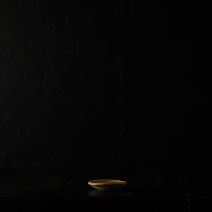 May include: A small, shallow, oval-shaped ceramic bowl with a light brown rim and interior sits on a dark surface. The background is a solid black, creating a high-contrast, minimalist composition. The bowl is centered.