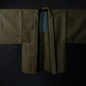 Peut inclure: Une veste haori vert olive avec des manches larges et un col structuré. Le vêtement présente un extérieur en tissu texturé uni et une doublure gris foncé contrastante visible au niveau du col intérieur, suspendu ouvert sur une barre.