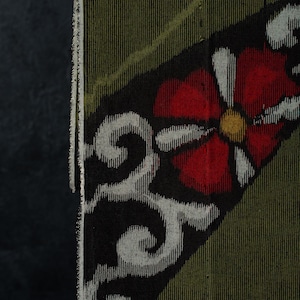 May include: A textile fabric featuring a dark olive green base with a prominent diagonal band in black. The black section displays a stylized red flower with a yellow center and white petals, accompanied by intricate white scrollwork patterns.