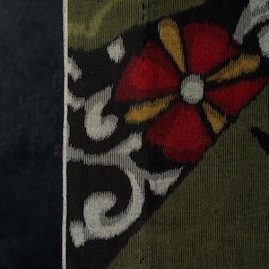 Puede incluir: Una tela textil con un fondo verde oliva oscuro y una sección triangular negra prominente. Esta sección muestra una flor roja estilizada con pétalos amarillos y un centro blanco, acompañada de intrincados patrones de volutas de color blanco.