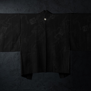 Puede incluir: Una chaqueta haori negra con un sutil patrón jacquard tonal de motivos florales y geométricos. La prenda tiene mangas anchas, una abertura frontal recta y un cierre circular único en el escote, exhibida sobre una barra horizontal.