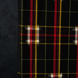 May include: A black fabric featuring a plaid pattern with intersecting red and yellow lines. The design includes small, rectangular, woven-in blocks of white, red, and yellow, creating a textured, grid-like appearance across the dark surface.