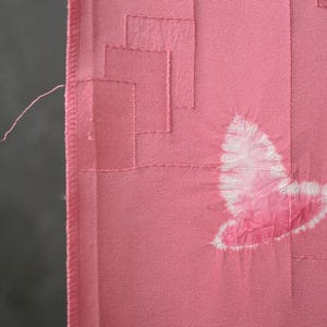 May include: Pink fabric with a geometric design and a butterfly motif. The fabric has a textured appearance with stitched details. The butterfly is a lighter shade of pink with white accents.
