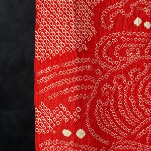 May include: A vibrant red fabric featuring a dense, intricate pattern of small, white, circular tie-dye resist marks. The textile displays a textured, crinkled surface with repeating geometric and swirling motifs created by the shibori technique.