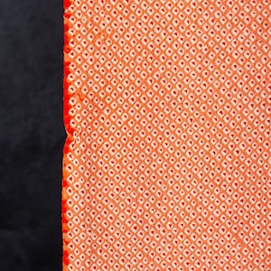 May include: A vibrant orange fabric featuring a dense, repeating pattern of small white squares arranged in a grid. The material has a textured surface with a distinct, straight vertical edge on the left side, showcasing the intricate weave.