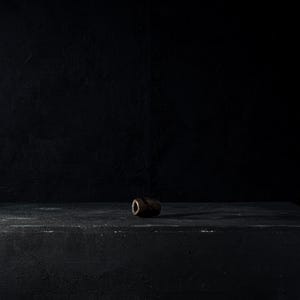 May include: A dark brown, cylindrical object with a hole through the center rests on a dark, textured surface. The object is centered on a black, rectangular block against a black background. The lighting creates shadows and highlights the object's shape.