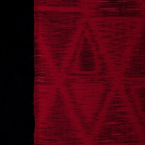 Pode incluir: Um tecido vermelho profundo com um padrão ikat escuro e subtil em forma de diamante. O material apresenta uma textura fibrosa horizontal em toda a trama, com uma borda vertical distinta e ligeiramente desfiada sobre um fundo preto.