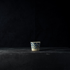 Può includere: Una piccola tazza in ceramica bianco sporco con un motivo geometrico blu. La tazza è cilindrica e poggia su una superficie scura e strutturata su uno sfondo nero. Il design presenta forme blu ripetute.