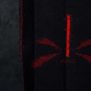 Op de afbeelding: Een zwarte stof met een geweven patroon van abstracte rode vormen. Het textiel heeft een zichtbare verticale rand met een gerafelde, getextureerde naad. De rode designelementen lijken enigszins wazig, wat een zacht en diffuus effect creëert.